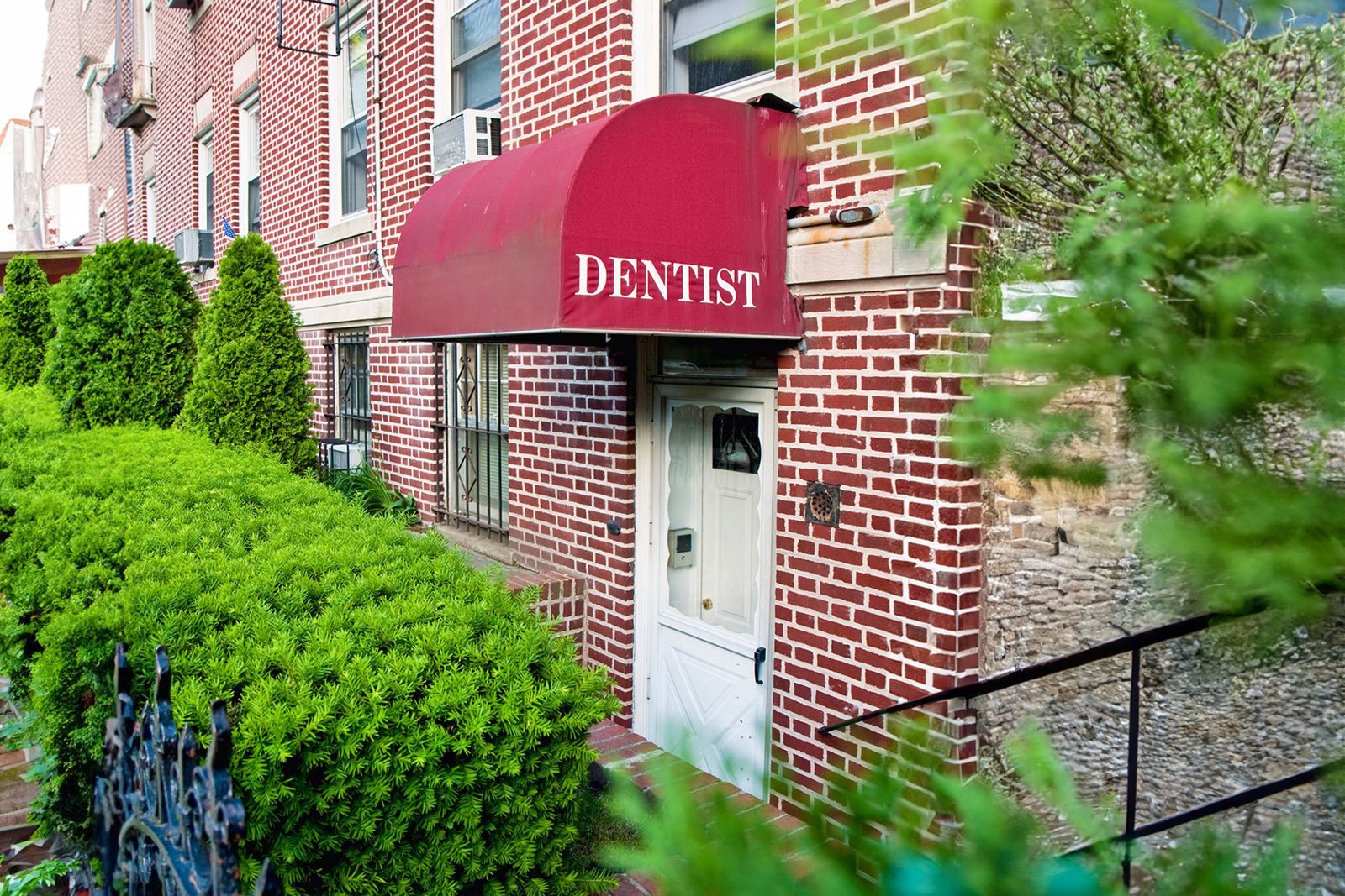 Entrance to a brick building with a red awning labeled Dentist, surrounded by green bushes and trees.