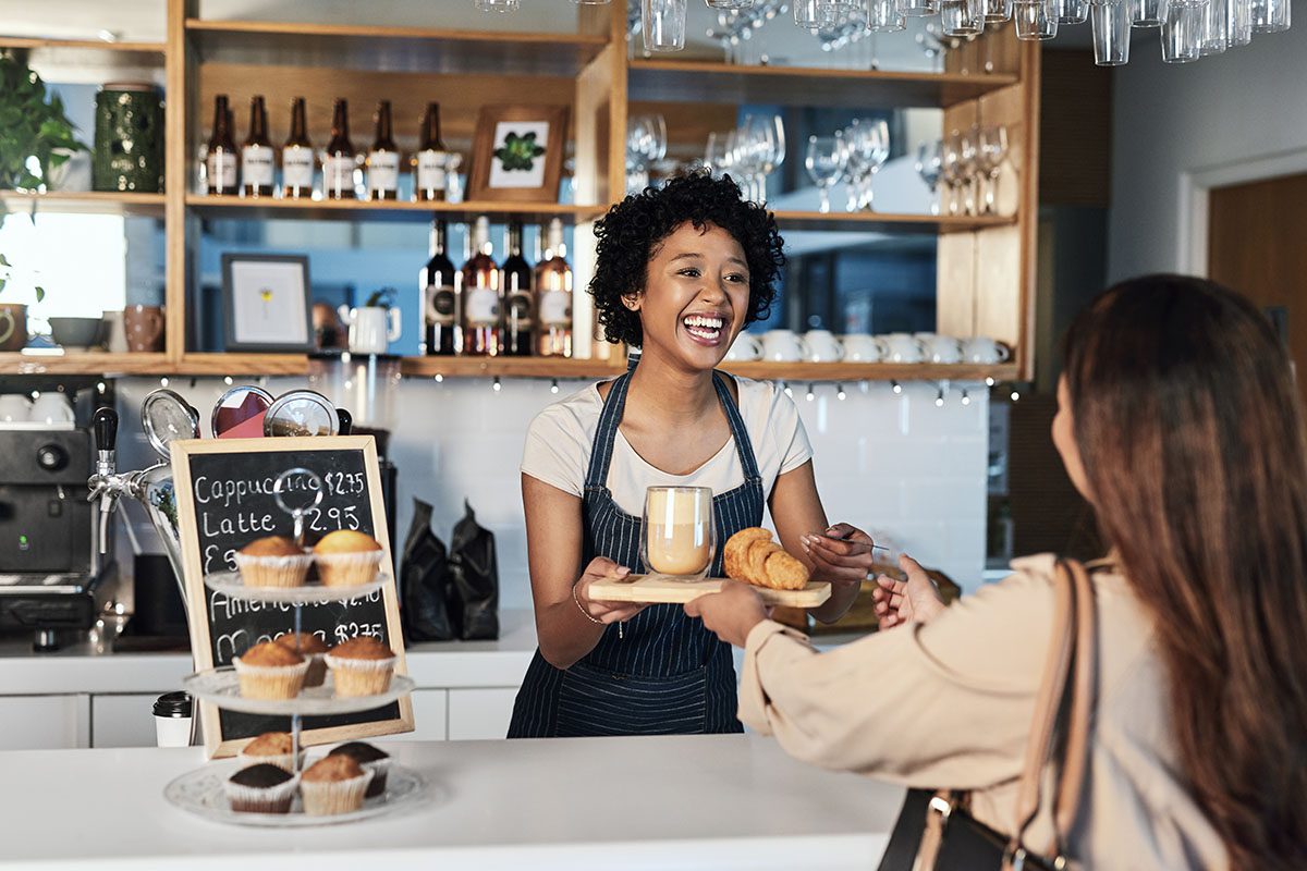 A barista behind a café counter hands a tray with a croissant and a drink to a customer; a chalkboard menu and pastries are visible on the counter.