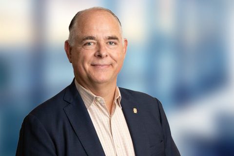 A middle-aged man in a suit jacket and light-colored shirt smiles at the camera against a blurred blue background.