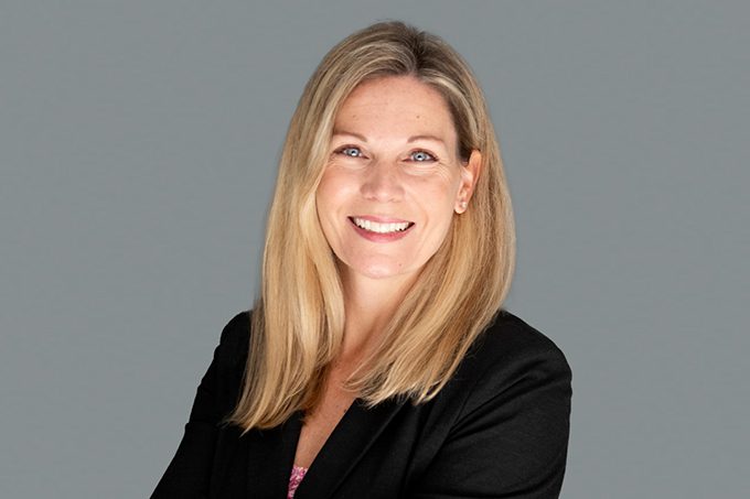 A woman with straight blonde hair, wearing a black blazer, smiles at the camera against a plain gray background.