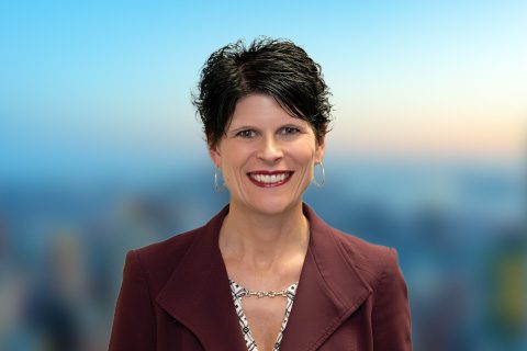 Dark haired woman in a blazer smiles in front of a bright blurred background