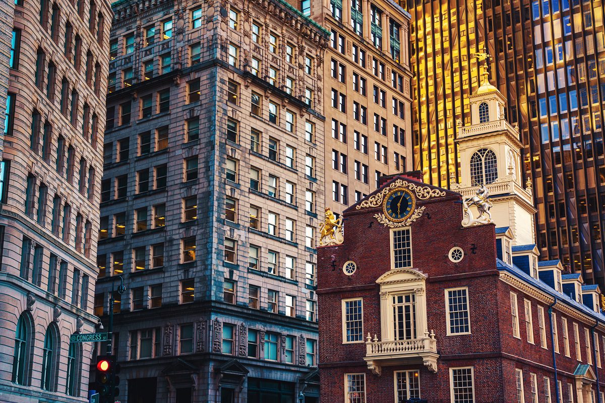Several tall office buildings surround a historic brick building with a clock and golden decorations on its facade, located at a city intersection.