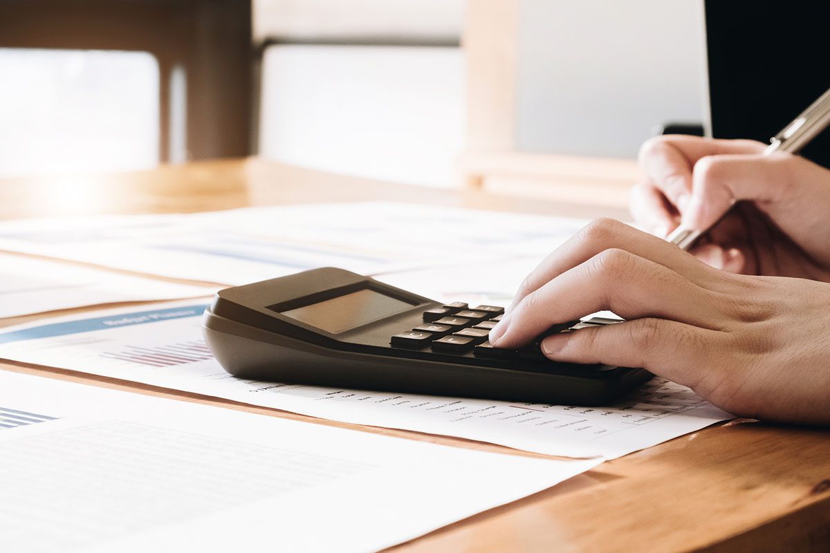 A person uses a calculator and writes on documents spread out on a wooden desk, with financial charts visible on the papers.