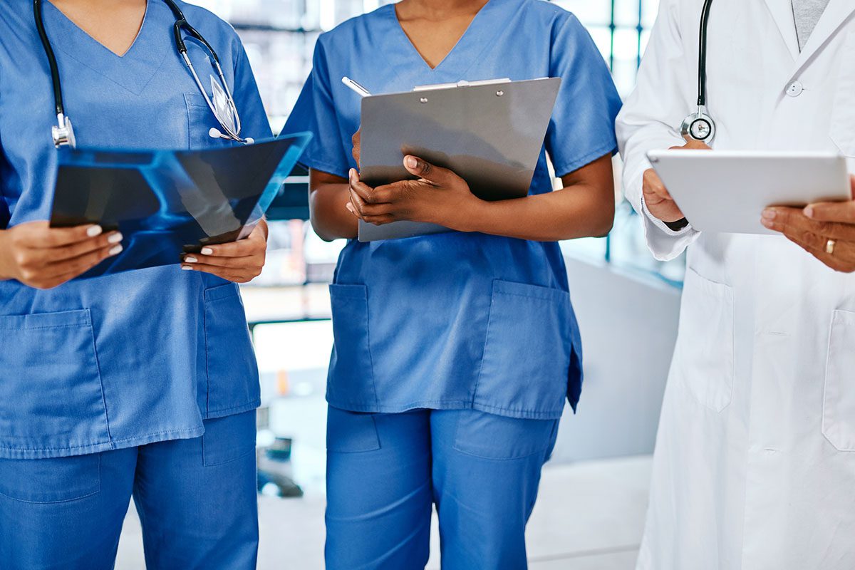 Three healthcare professionals, two in blue scrubs holding medical charts and one in a white coat holding a tablet, stand together indoors.
