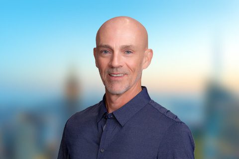 A middle-aged man with a shaved head and a short beard, wearing a dark blue button-up shirt, stands in front of a blurred cityscape background.