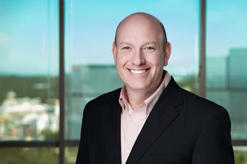 Bald man in a black blazer and light pink shirt smiling, standing indoors with large windows and a cityscape in the background.