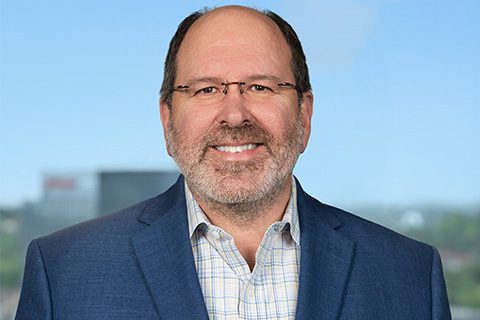 A middle-aged man with glasses, short hair, and a beard, wearing a blue blazer over a checkered shirt, smiles at the camera against a blurred outdoor background.