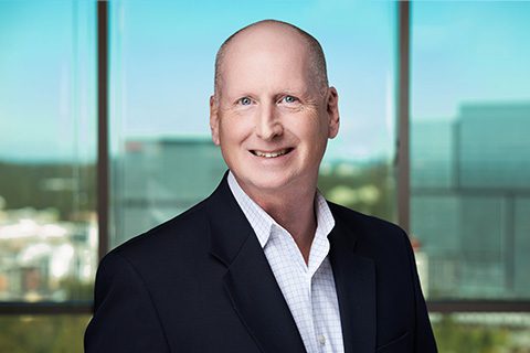 A bald man in a dark suit jacket and white shirt smiles in front of a window with an urban cityscape in the background.