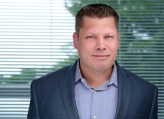A man in a blue checked blazer and striped shirt stands in front of horizontal blinds with greenery visible outside.