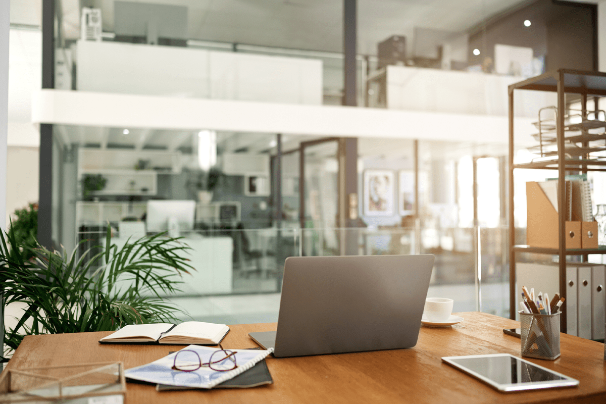 A modern office workspace with a laptop, notebook, papers, and pen on a wooden desk; glass walls and office furniture are visible in the background.