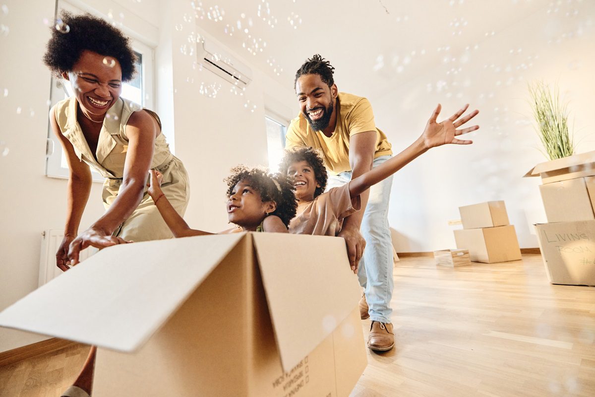 A family of four smiles and plays with cardboard boxes in a bright room with moving boxes, suggesting they have just moved into a new home.