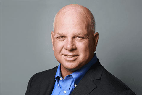 A middle-aged man with a bald head, wearing a blue shirt and dark blazer, poses against a gray background.