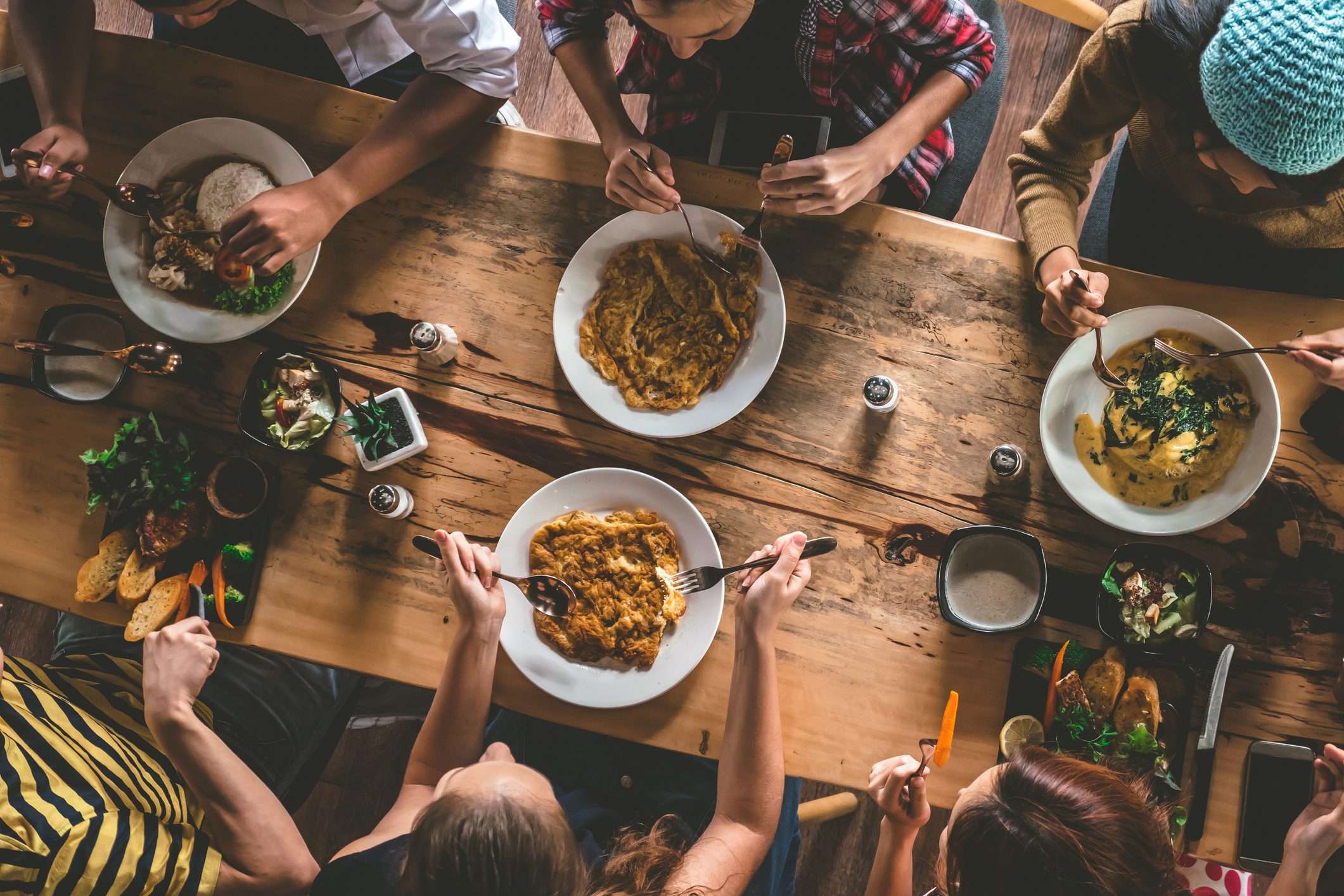 Five people sit around a wooden table eating meals from large bowls, with side dishes, bread, and drinks also on the table. The photo is taken from above.