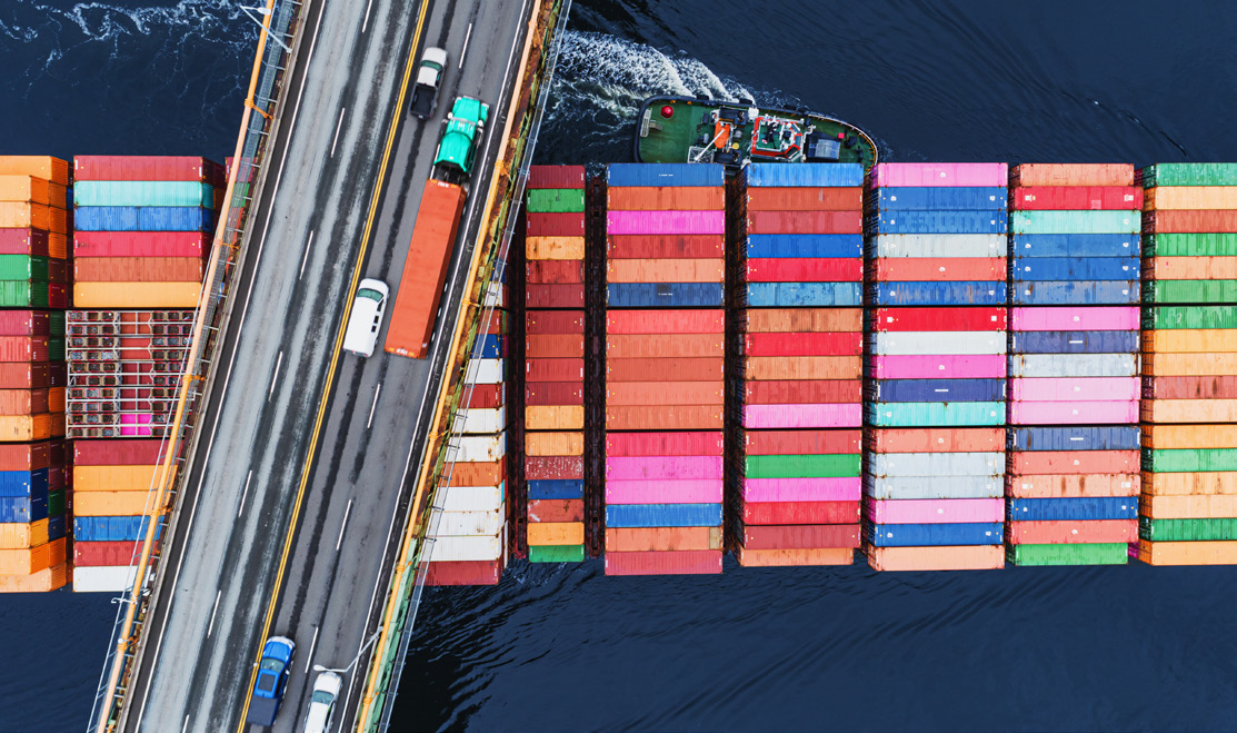 Aerial view of a cargo ship loaded with colorful shipping containers passing under a bridge with several vehicles.