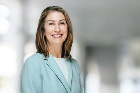 A middle-aged woman with shoulder-length brown hair wearing a light blue blazer smiles while standing in a bright, blurred indoor setting.