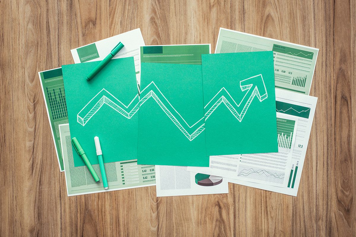 Several green papers and charts with financial graphs are spread on a wooden table. A rising arrow is drawn across three green sheets, symbolizing growth or upward trends.