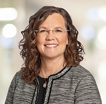 A woman with shoulder-length curly brown hair, wearing glasses and a gray jacket, smiles at the camera against a softly blurred background.