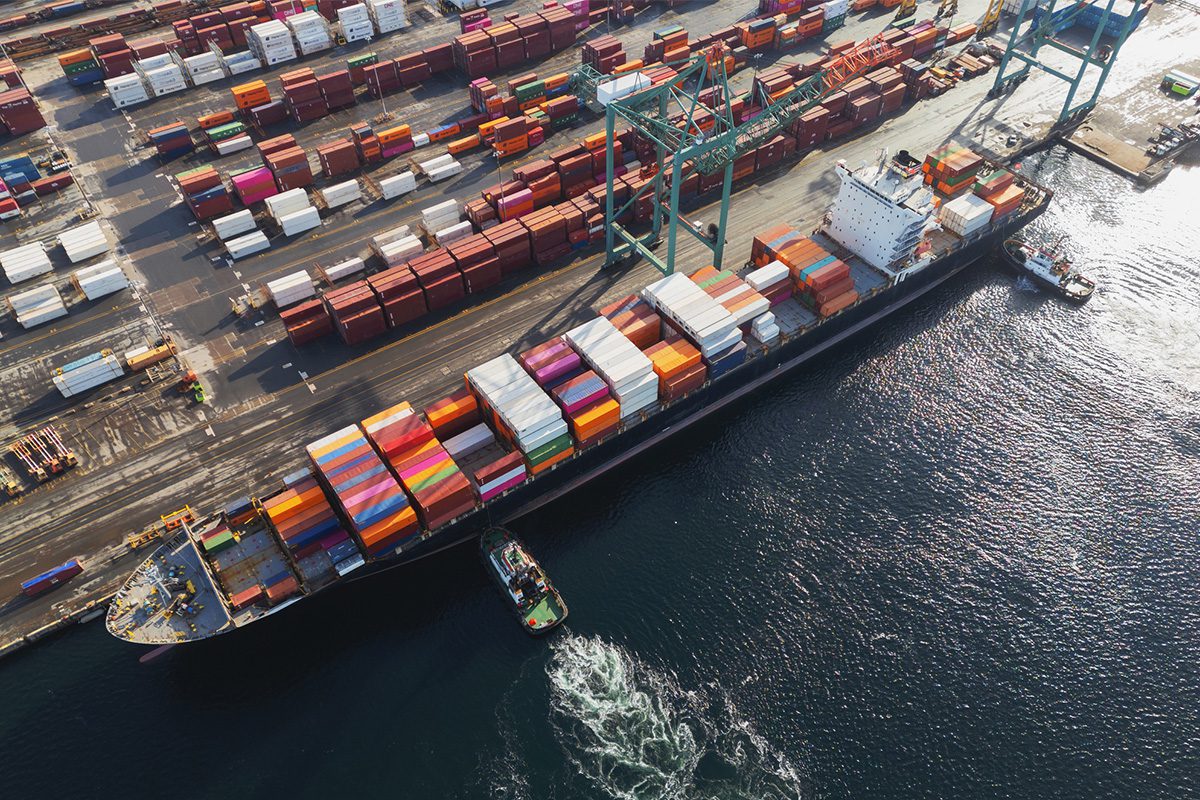 Aerial view of a cargo ship docked at a port, loaded with colorful shipping containers, with cranes and stacked containers nearby.