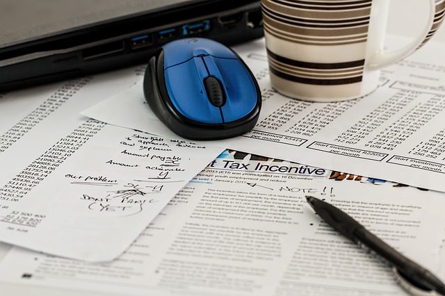 A blue computer mouse, a coffee mug, and a pen rest on documents, spreadsheets, and handwritten notes on a cluttered desk.