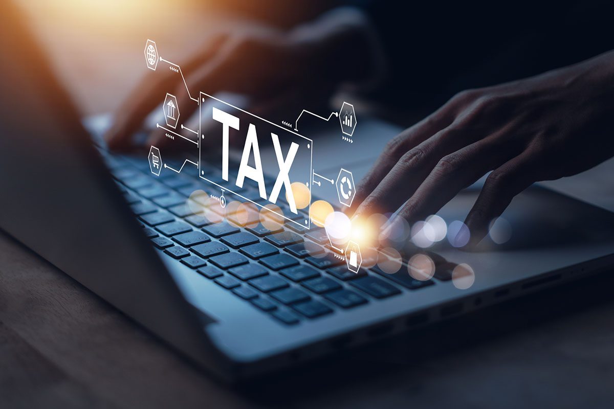 Hands typing on a laptop keyboard with a digital TAX graphic and related financial icons overlaid on the image.