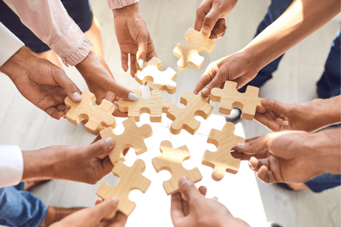 A group of people hold large wooden puzzle pieces together, forming a circle over a light-colored surface.