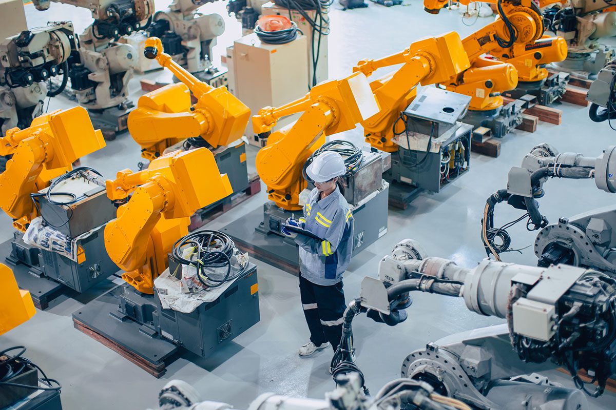 A worker in safety gear inspects large yellow industrial robotic arms in a factory or manufacturing facility.