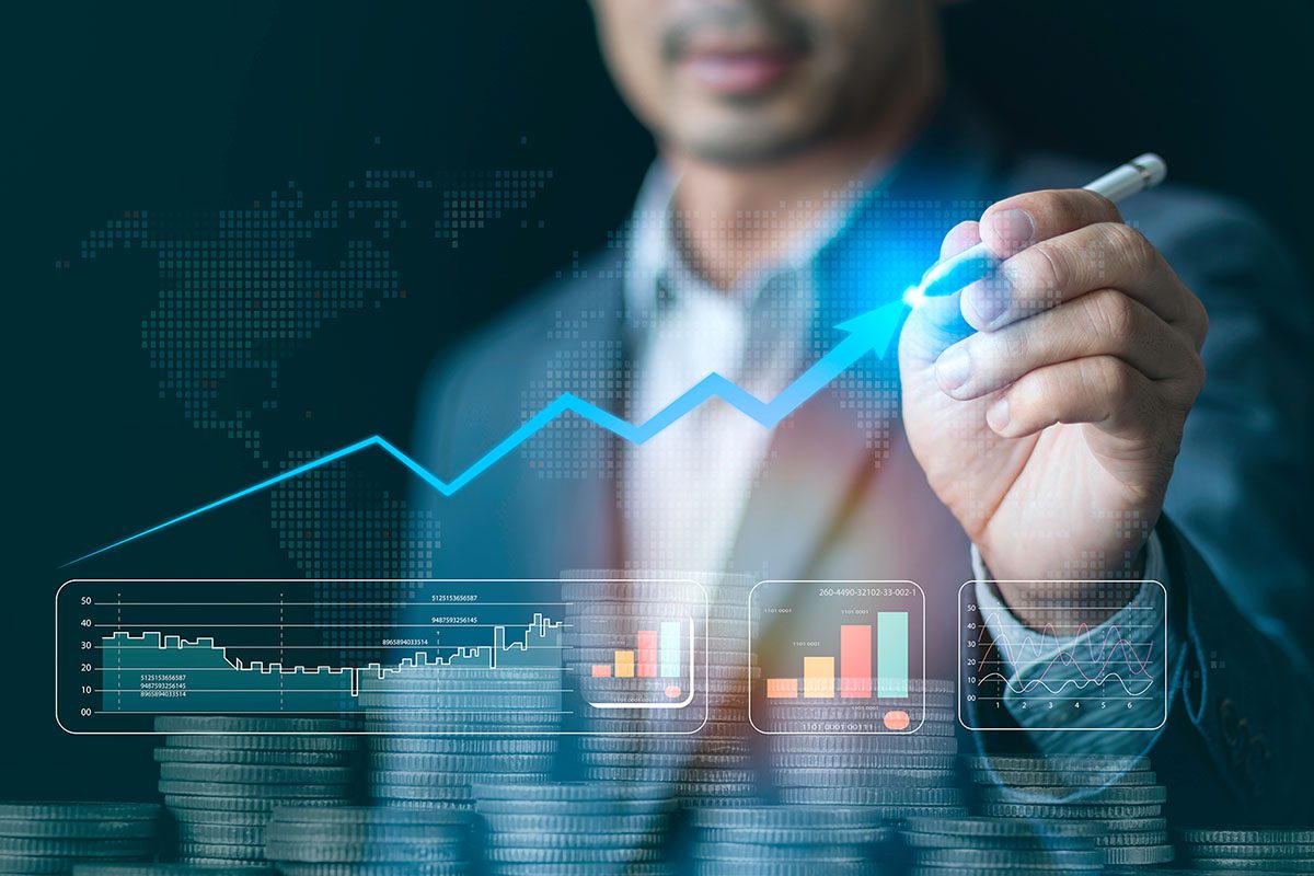 A person in a suit draws an upward trending arrow on a virtual screen displaying financial graphs and data, with stacks of coins in the foreground.