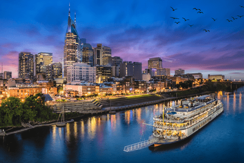 A riverboat docked on a river in front of a city skyline at dusk, with tall buildings illuminated and birds flying in the sky.