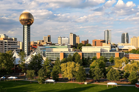 Cityscape of downtown Knoxville, Tennessee, featuring the Sunsphere, modern buildings, and a green park with scattered trees in the foreground.