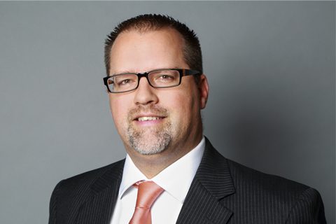 A man wearing glasses, a dark pinstripe suit, white shirt, and a peach-colored tie, posing against a plain gray background.