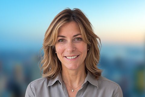 A woman with shoulder-length light brown hair, wearing a dark blouse, stands outdoors with a blurred cityscape in the background.