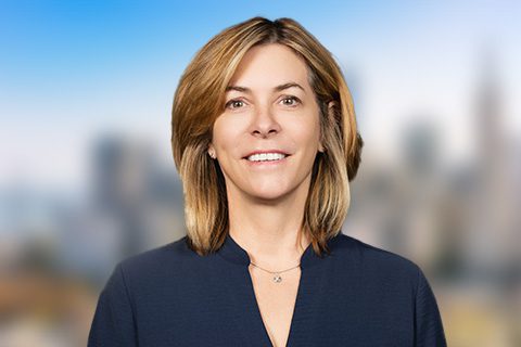 A woman with shoulder-length light brown hair, wearing a dark blouse, stands outdoors with a blurred cityscape in the background.