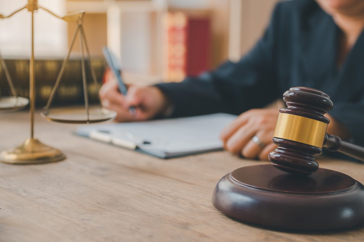 A judge writes on paperwork at a desk with a gavel and scales of justice in the foreground.