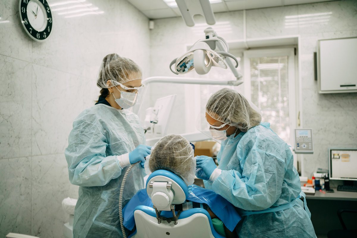 Two dental professionals in protective gear are performing a dental procedure on a patient seated in a dental chair in a clinic.