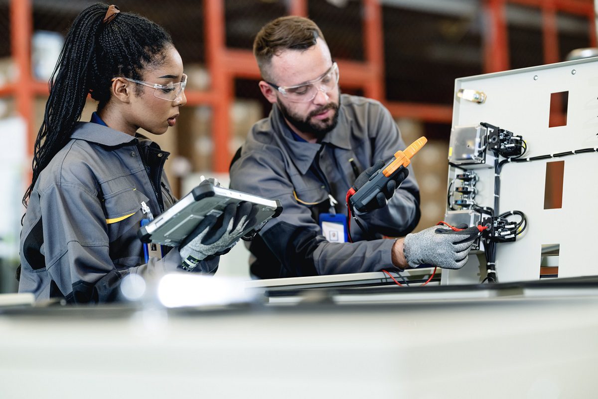 Two engineers in protective gear inspect and test electrical components on a control panel; one holds a tablet while the other uses testing equipment.