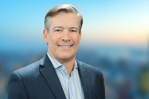 A man in a business suit smiles at the camera with a blurred cityscape background.