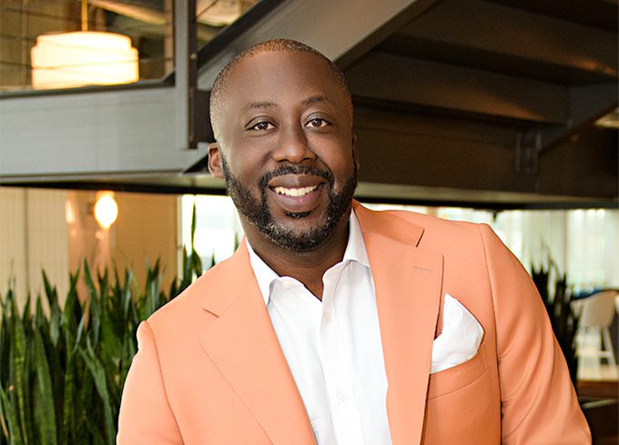 A man in a light orange suit jacket and white shirt stands indoors, smiling, with green plants and modern lighting in the background.
