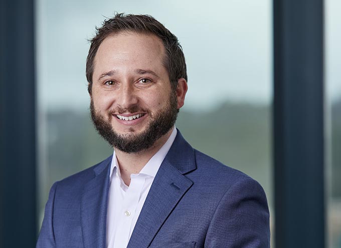 A man with a beard and short brown hair, wearing a blue suit jacket and white shirt, standing in front of a window.