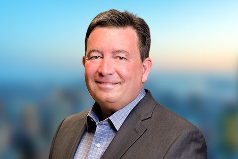 Dark haired man in a gray blazer smiles in front of a bright window with a blurred city view.