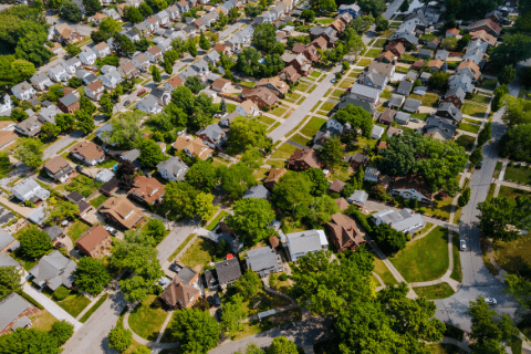 Aerial view of a suburban neighborhood with tree-lined streets, single-family houses, lawns, and intersecting roads.