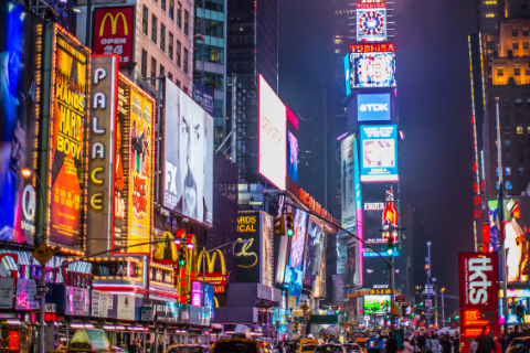 Crowded street in Times Square, New York City, at night with bright neon billboards, advertisements, and yellow taxis visible.