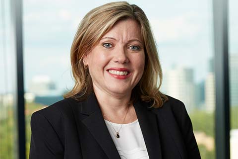 A woman with shoulder-length blonde hair, wearing a black blazer and white top, smiles in front of a window with a cityscape background.