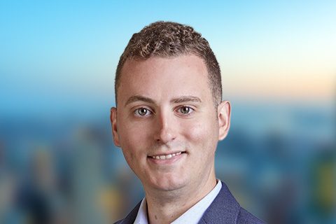 Dark, curly-haired man smiles in front of a bright window with a blurred city view