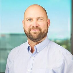 A bald man with a beard wearing a light blue button-up shirt stands in front of a window with a blurred outdoor background.