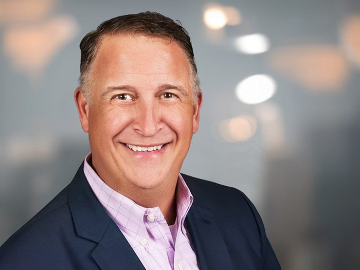 Smiling man in a suit jacket and light purple shirt poses in front of a blurred background with lights.