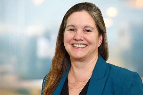 A woman with long brown hair, wearing a teal blazer and black top, smiles at the camera against a blurred indoor background.