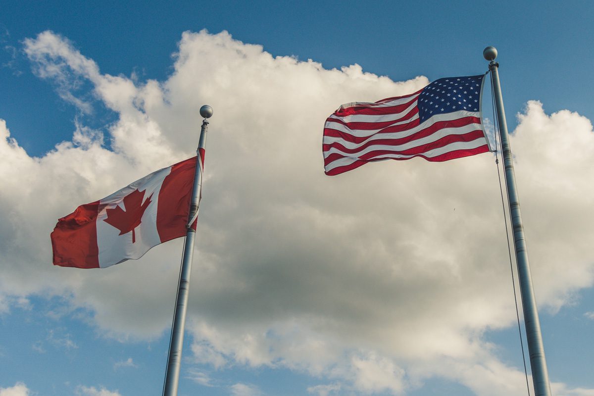 The Canadian flag and the United States flag are flying on separate flagpoles against a partly cloudy sky.