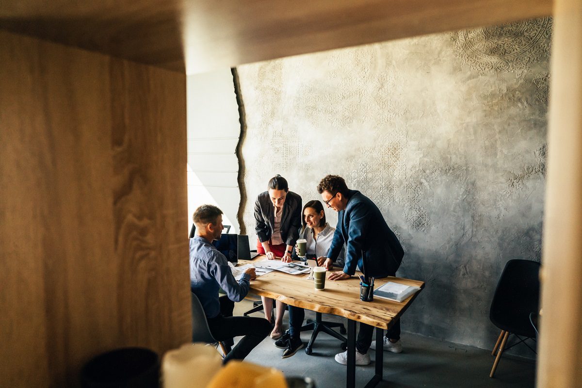 Four people sit and stand around a wooden table in an office, looking at documents and laptops, viewed through a shelf.