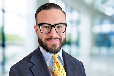 Dark haired, bearded man in glasses and a blazer smiles in a blurred bright lobby