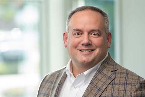 Man in a plaid blazer and white shirt smiles at the camera, standing indoors with blurred windows in the background.